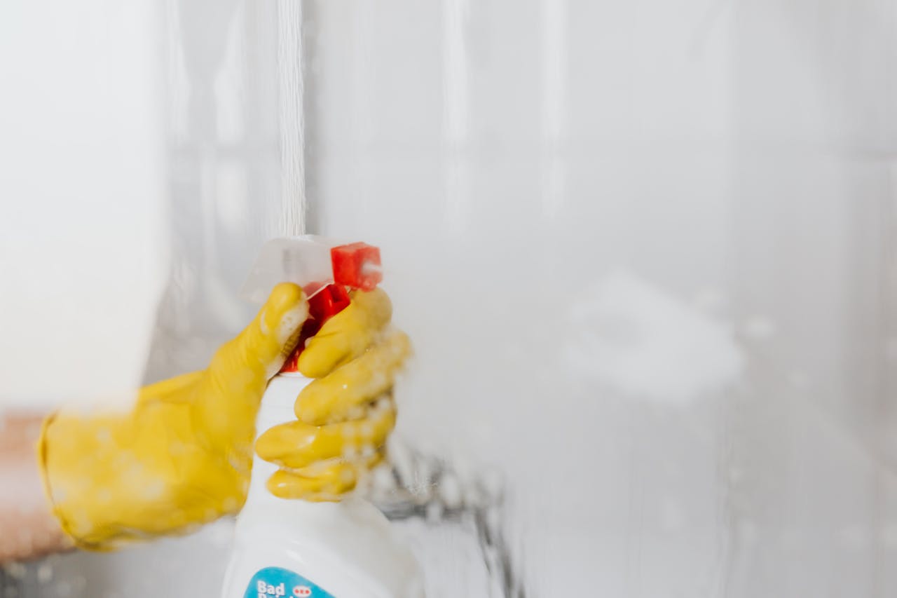services-05 Crop unrecognizable person in yellow latex protective glove spraying detergent from bottle with plastic dispenser at glass of shower cabin